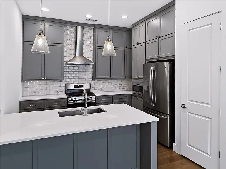 Kitchen featuring an island with an undermount sink, quartz countertops, gray shaker-style cabinetry, stainless steel appliances, and a subway tile backsplash