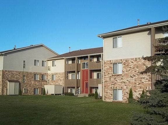 Brown brick and brown sided building with wooden balconies on the second and third floor. There is an expansive green lawn and a pine tree in the right of the photo.