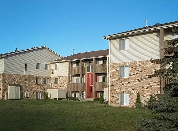 Brown brick and brown sided building with wooden balconies on the second and third floor. There is an expansive green lawn and a pine tree in the right of the photo. - Brighton Cove