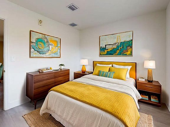 Inviting bedroom with modern furnishings, featuring a cozy bed with vibrant yellow and teal accents, elegant wooden furniture, and abstract art for a refreshing ambiance.