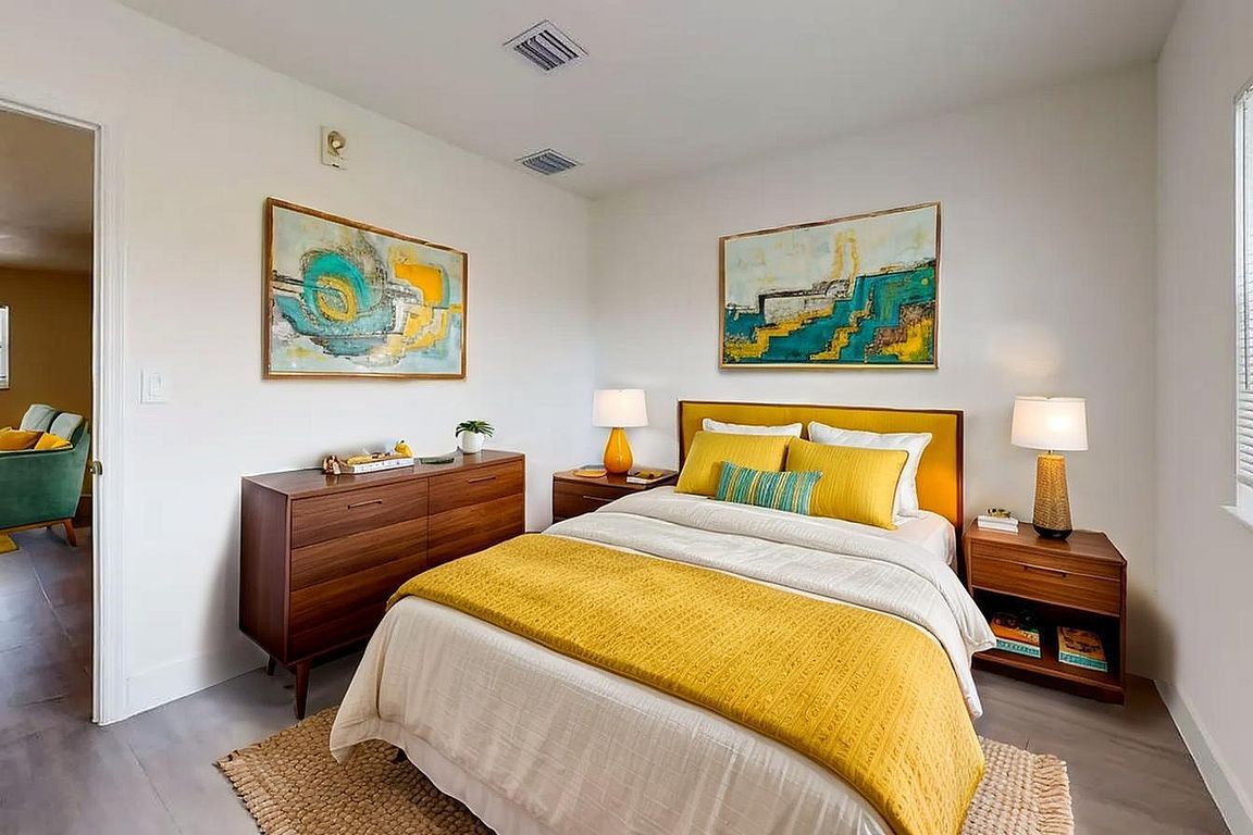 Inviting bedroom with modern furnishings, featuring a cozy bed with vibrant yellow and teal accents, elegant wooden furniture, and abstract art for a refreshing ambiance.