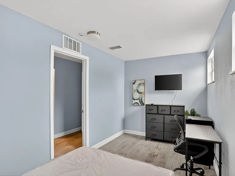Bedroom includes a work desk, dresser, and wall-mounted TV, with large windows and durable flooring for easy upkeep.