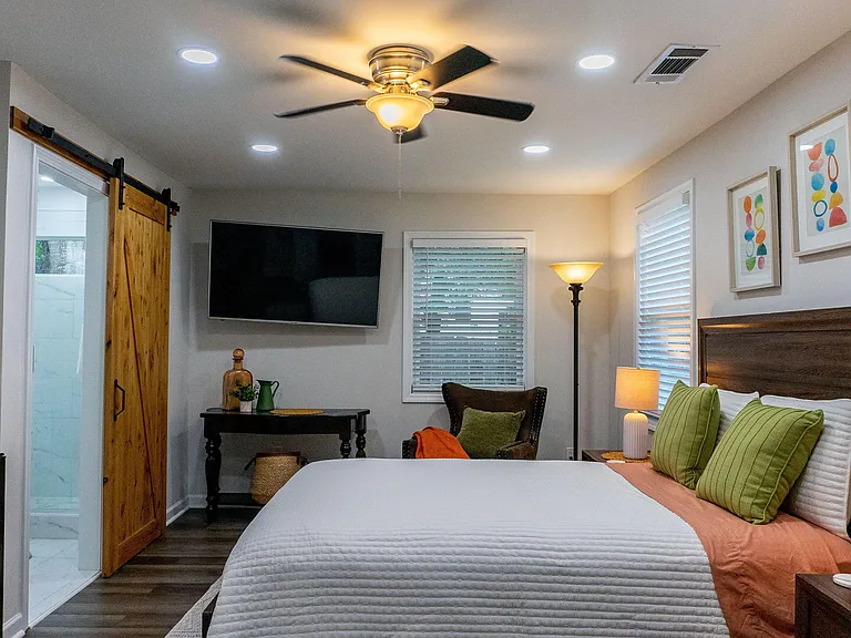 Furnished bedroom with ceiling fan, wall-mounted TV, and windows with blinds, plus a sliding barn door to the bath.