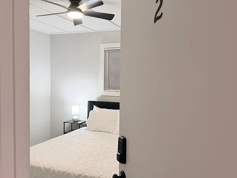 Private bedroom with a comfortable bed, ceiling fan, and bedside table with lamp, plus a window with blinds.