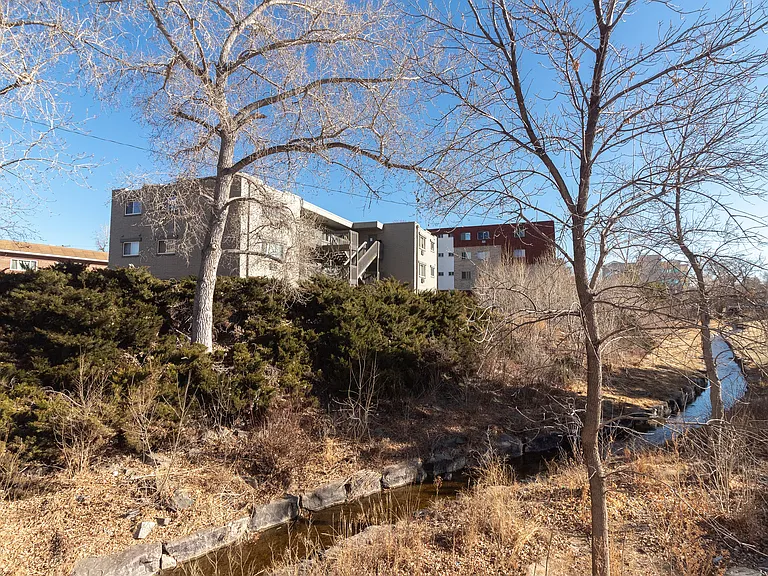 Charming apartment complex nestled among lush greenery with a serene creek and expansive blue sky view.