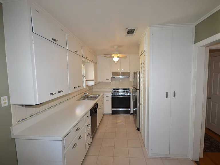 Galley kitchen featuring extensive white cabinetry, light-toned countertops, a double-basin stainless steel sink, tile flooring, and integrated appliances including a range and refrigerator