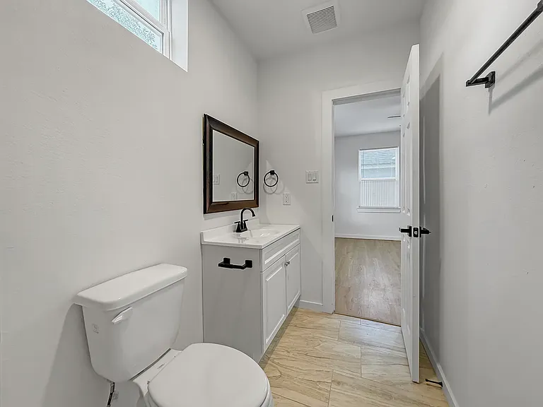 This photo shows a bright, modern bathroom with a white vanity, a sleek black faucet, and a framed mirror. The space features a toilet and light wood flooring, with a small window providing natural light. A door leads to another room, enhancing accessibil