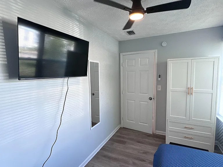 Private bedroom with wood-look flooring, ceiling fan, wall-mounted TV, and a wardrobe with drawers for added storage.