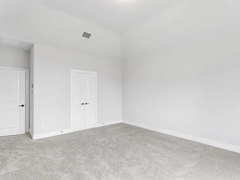 Spacious interior room featuring a vaulted ceiling, light gray carpeting, crisp white walls, a single panel door with black hardware, and a bi-fold door with black hardware
