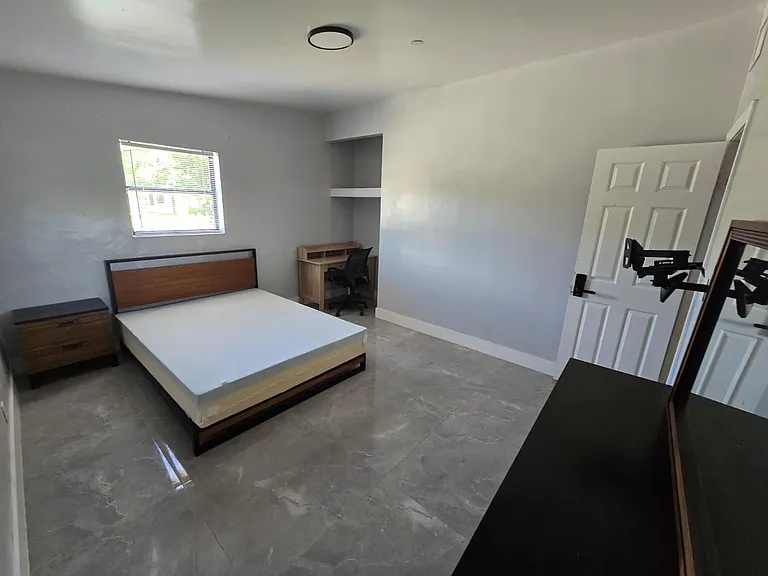 Furnished bedroom with a full bed, nightstand, and a dedicated desk area, plus tile flooring and a window with blinds.