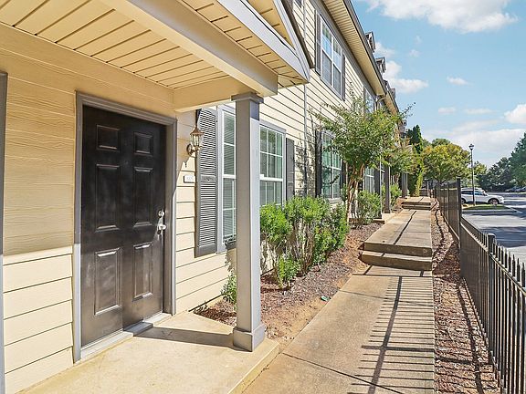 Charming apartment entrance with black door, surrounded by lush greenery and well-maintained pathway.