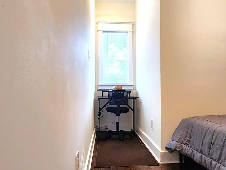 Narrow bedroom with wood-look flooring, a dedicated desk nook by the window, and overhead lighting for everyday use.