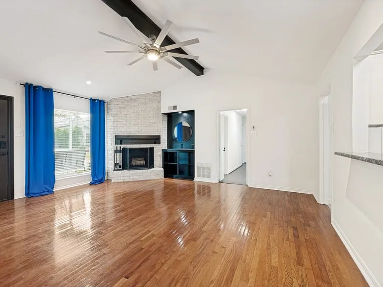 Spacious living area featuring wood-finish flooring, a white brick fireplace, vaulted ceilings with dark wood beams, and a modern ceiling fan