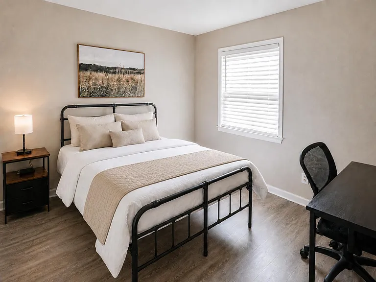 Furnished bedroom with a full-size bed, nightstand with lamp, and a dedicated desk workspace beside the window.