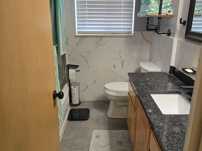 Full bathroom with tub/shower combo, tiled walls, ample vanity counter space, and open shelving for linens.