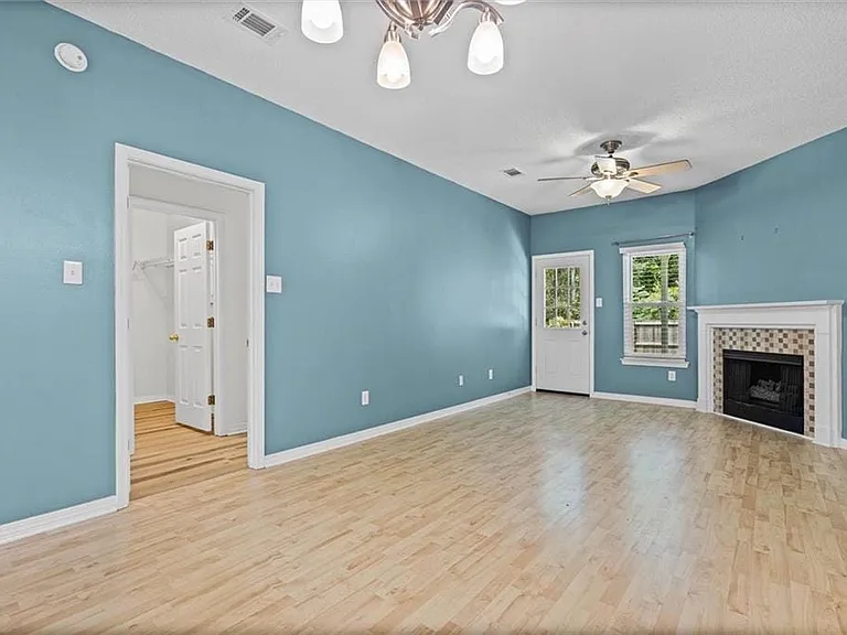Spacious room featuring wood-finish flooring, a fireplace with a tiled surround and white mantel, a ceiling fan, and a light-filled exterior door with an adjacent window