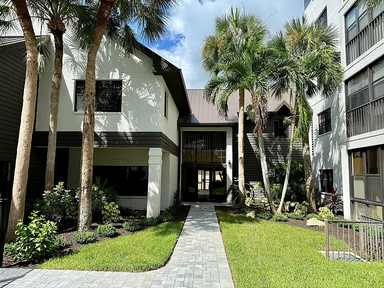 Charming apartment entrance surrounded by lush green landscaping and palm trees, featuring modern architecture and tranquil vibes.