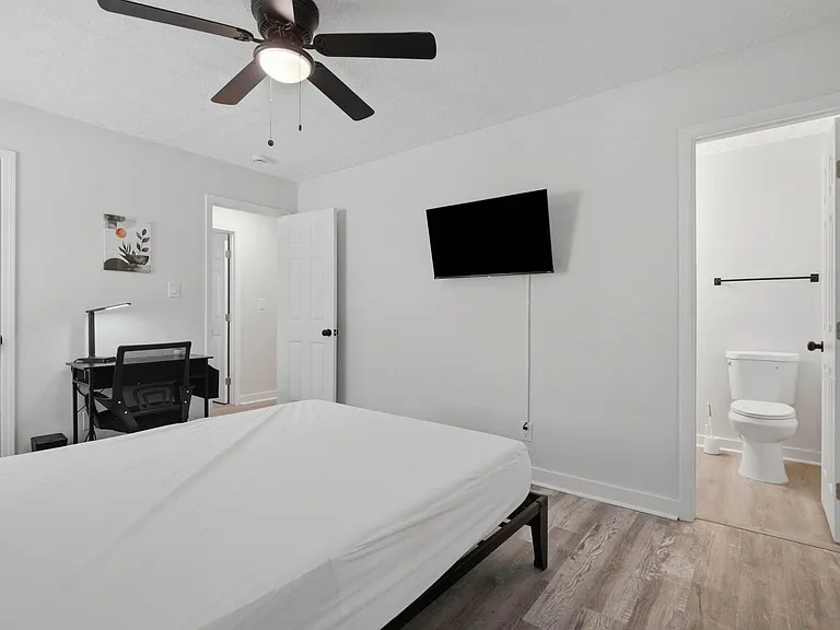 Furnished bedroom with ceiling fan, work desk, and wall-mounted TV, plus convenient access to a nearby bathroom.