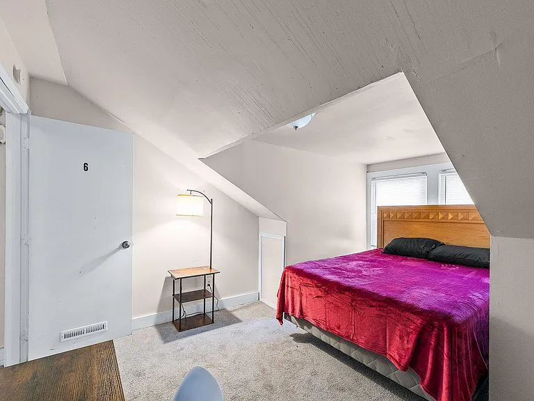 Carpeted bedroom with a full-size bed, bedside table and lamp, plus a window with blinds for natural light.