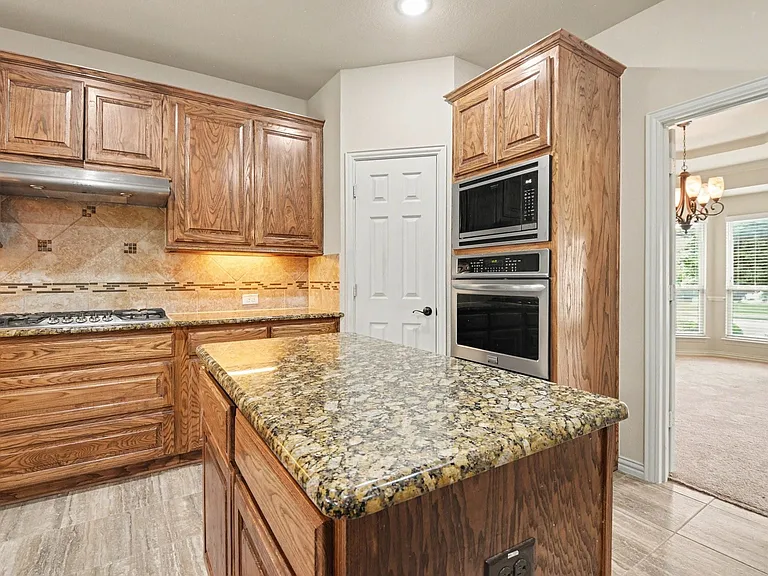 Kitchen featuring a central island with a granite countertop, wood cabinetry, built-in stainless steel appliances, and a gas cooktop with a tile backsplash