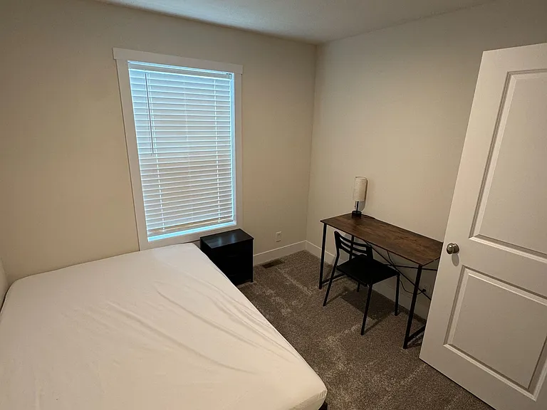 Private bedroom with a large window and blinds, carpet flooring, and a dedicated desk area for work or study.