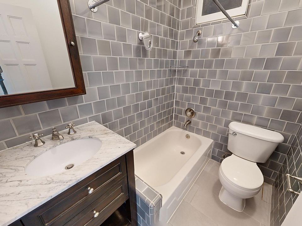 Compact bathroom with tiled walls, featuring a bathtub, toilet, and a sink counter. Warm wooden-framed mirror adds a classic touch.