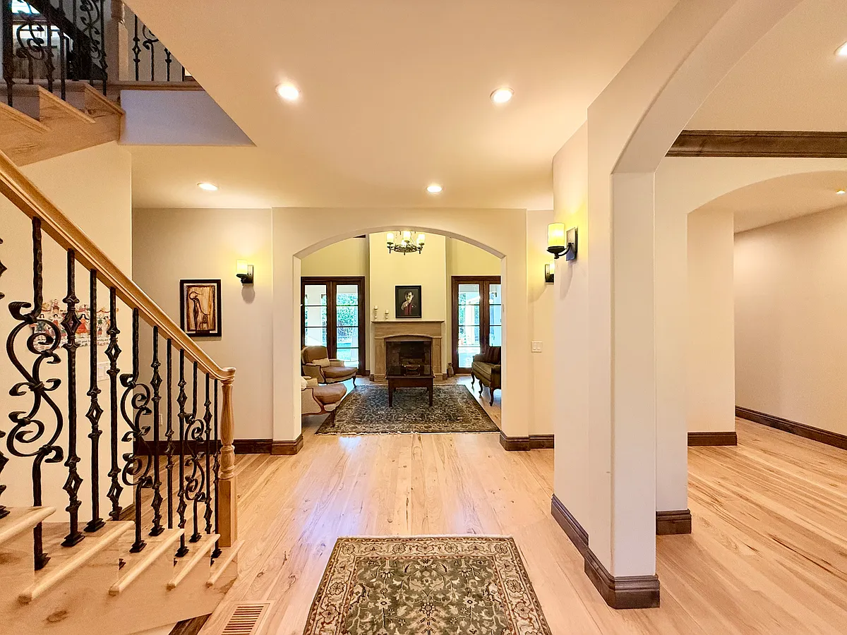 Inviting foyer with wide-plank hardwood floors, elegant staircase with wrought-iron balusters, and soft recessed lighting. Arched openings lead to a formal living area with fireplace, creating a bright, open entry that sets a warm, refined tone - Cowper St