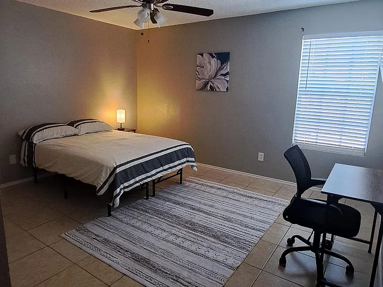 Spacious bedroom with ceiling fan, tiled floors, a large window with blinds, and room for a desk and seating area.