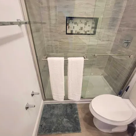 Modern tiled shower with glass door, bath mat, towels, and towel racks available.