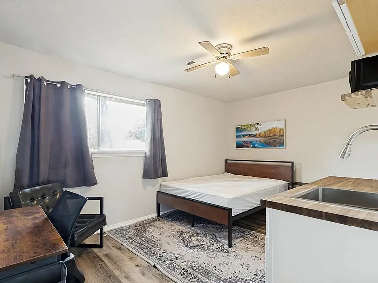 Furnished bedroom with a bed, desk area, and seating, plus a TV, ceiling fan and window with curtains for comfortable living.
