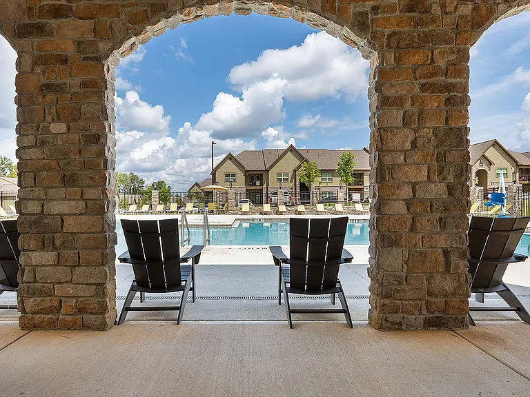 Inviting view of a luxurious apartment community pool framed by elegant stone arches, featuring comfortable lounge chairs under a bright blue sky.