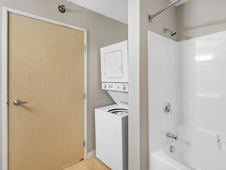 Modern bathroom with in-unit laundry featuring a stacked washer and dryer beside a sleek bathtub and neutral-toned door. Modern bathroom with in-unit laundry featuring a stacked washer and dryer beside a sleek bathtub and neutral-toned door.