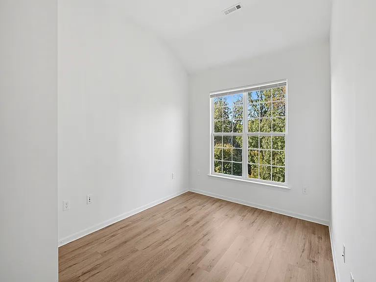 Bright and airy room with large window, elegant wooden flooring, and fresh white walls, offering a serene view of lush trees.