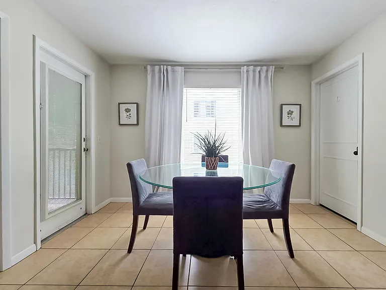 Dining area with a round glass-top table, seating for four, tiled floors, and a large window with curtain panels.