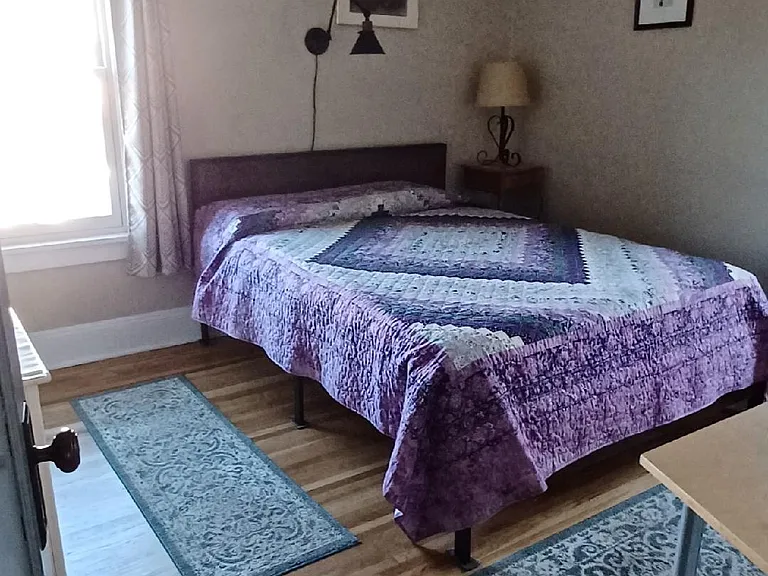 This bedroom has 2 windows with room darkening shade and drapes, bedside table, shelves with decorative storage baskets (or dresser) and a TV/work table.