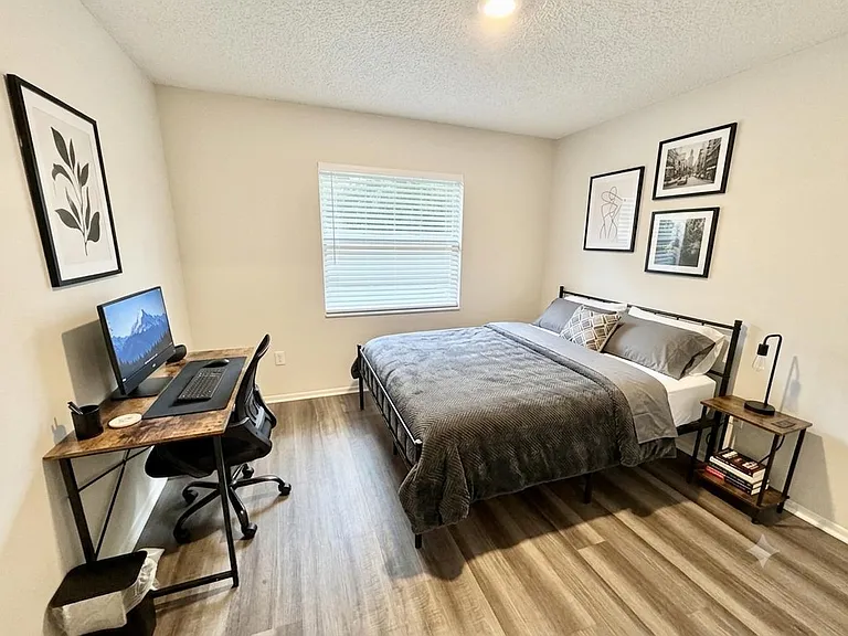 Furnished bedroom with a full bed, work desk, and nightstand, plus a window with blinds and durable wood-look floors.