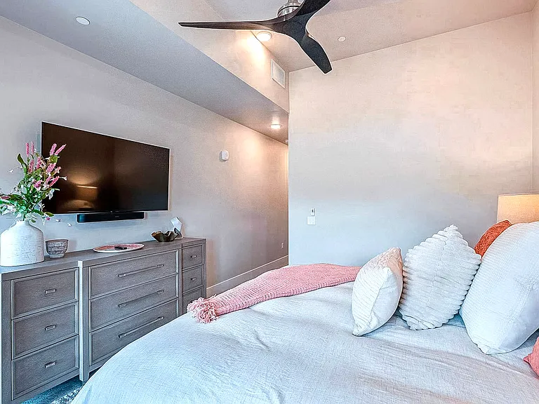 Third bedroom with wall-mounted TV, dresser, and soft pink and white bedding in a calm, neutral color palette