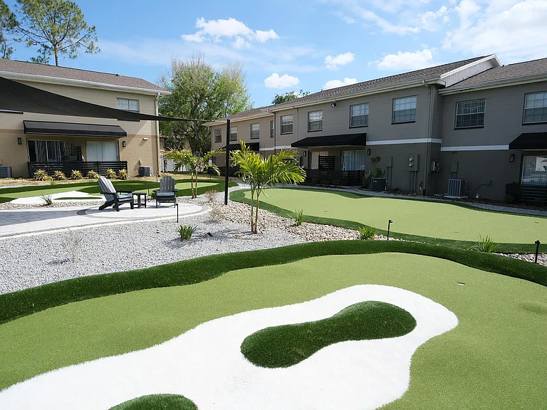 Inviting apartment courtyard with lush green mini-golf course, comfortable outdoor seating, and modern two-story buildings.