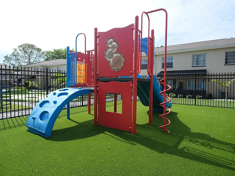 Vibrant playground with colorful slides and climbing structures, surrounded by lush greenery and secure fencing in a family-friendly community.