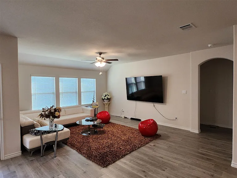Spacious living area featuring wood-finish flooring, a ceiling fan with integrated lighting, and multiple windows with blinds