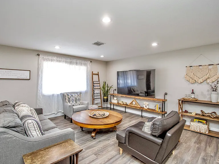 Cozy, inviting living room at Casa Del Sol, Tempe rental home. Features a large 80" smart TV, ample seating with a gray pullout sofa and accent chairs, boho wall decor, vintage round coffee table. Ideal space for relaxation in a top Tempe, AZ rental.