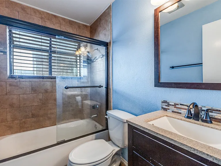 Bathroom featuring a tiled shower/tub combination with glass enclosure, window with blinds, vanity with integrated sink and mosaic tile backsplash, and dark wood-frame mirror