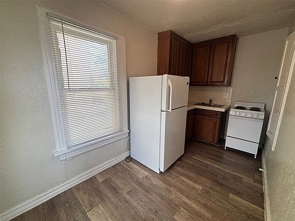 Compact kitchen space featuring wood-finish flooring, a double-hung window with blinds, and white baseboard trim