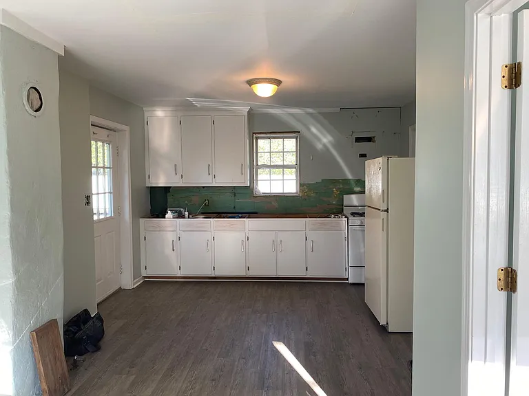 Partially renovated kitchen area with white cabinets. Entry door to the left. Completed kitchen will have white tile backsplash and grey granite countertops. Gas stove and oven.