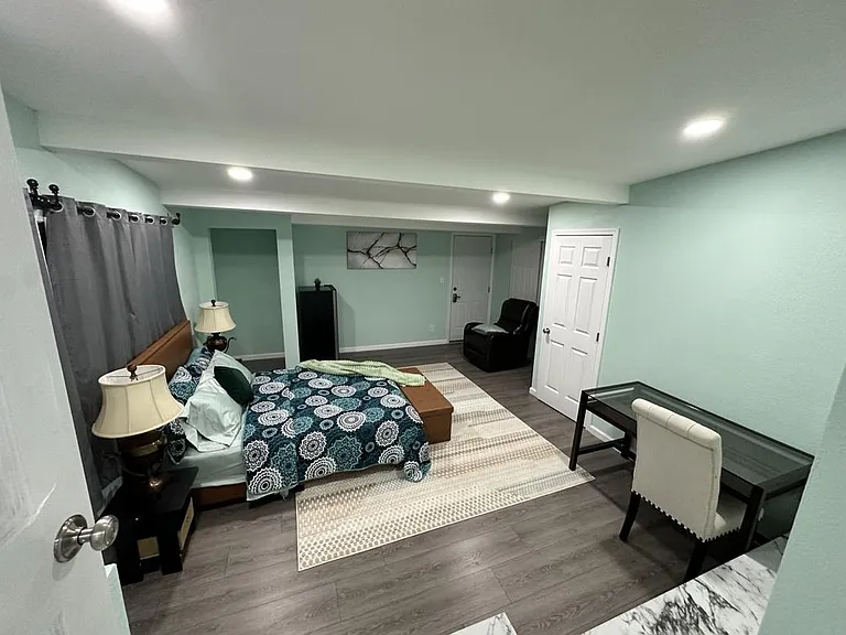 Spacious furnished bedroom with a queen bed, nightstands, a desk workspace, and a sitting chair on durable flooring.