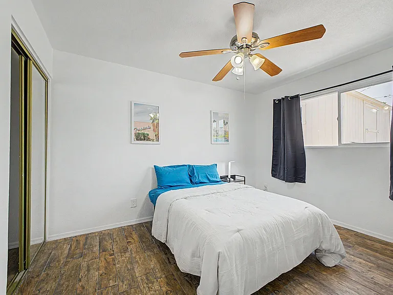 Private bedroom with wood-look flooring, mirrored closet doors, and a ceiling fan with light; window with curtains.