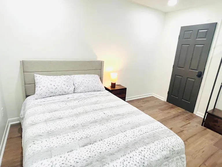 Furnished bedroom with a full bed, nightstand lamp, wood-style flooring, and an open clothes rack for easy storage.