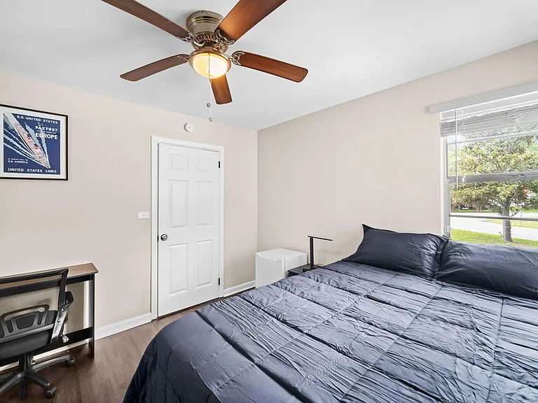 Furnished bedroom with a full bed, bedside storage, ceiling fan, and a workspace, plus a large window with neighborhood views