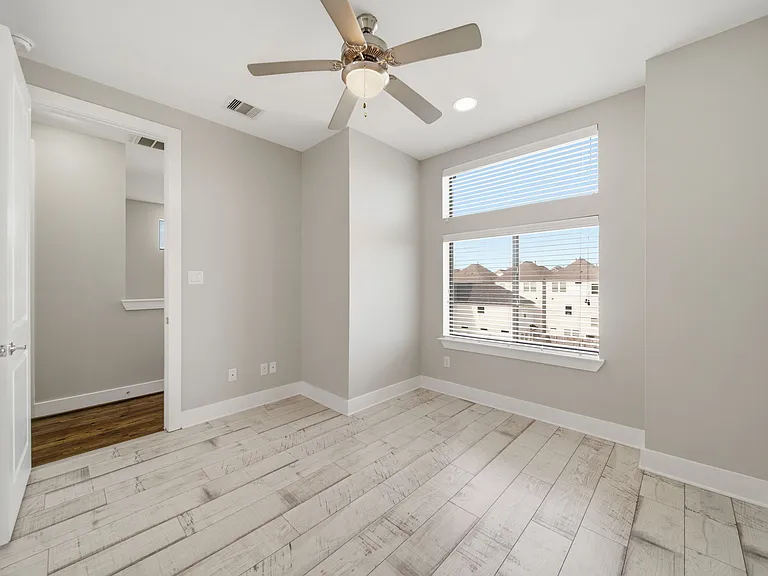 Bright, modern third-floor bedroom featuring light wood flooring, neutral finishes, and a ceiling fan. Large windows with blinds provide excellent natural light, creating an airy, inviting space.