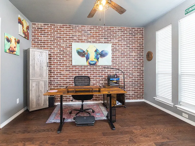 Dedicated office space with a desk, ergonomic chair, and coffee cabinet. Brick accent wall and ceiling fan create a cozy and inspiring work environment.